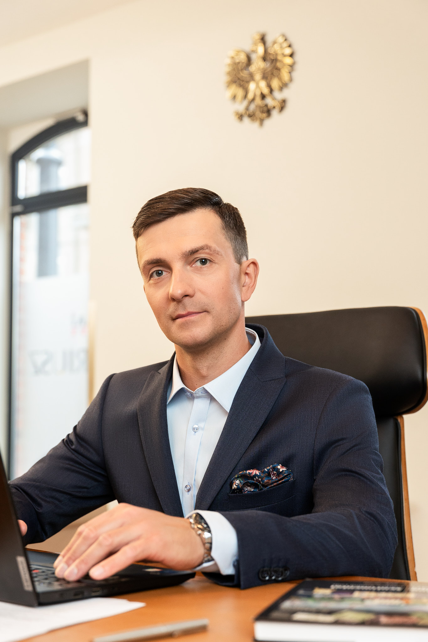 Notariusz Aleksander Wasiak Zdjęcie siedzącego przy biurku na czarnym fotelu Notariusza Aleksander Wasiaka. Ubrany jest w granatowy garnitur i niebieską koszulę. Trzyma dłonie na otwartym laptopie. W tle na ścianie widać zawieszone złote godło polskie w postaci orła.