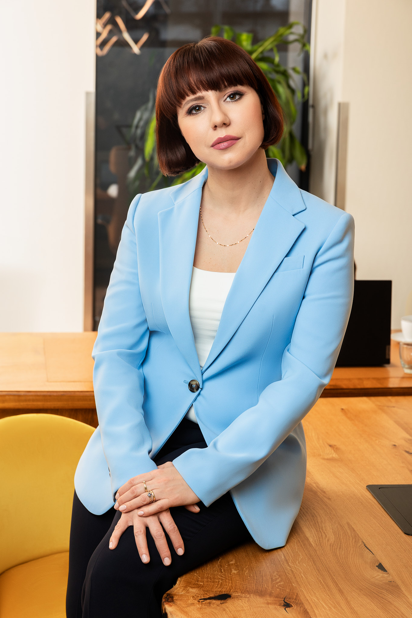 Notariusz Magdalena Kułaj Zdjęcie siedzącej na biurku Notariusz Magdaleny Kułaj, ubranej w niebieską marynarkę i białą koszulkę oraz czarne spodnie. Trzyma na nich złożone ręce. W tle widać dużego kwiatka oraz witrynę.