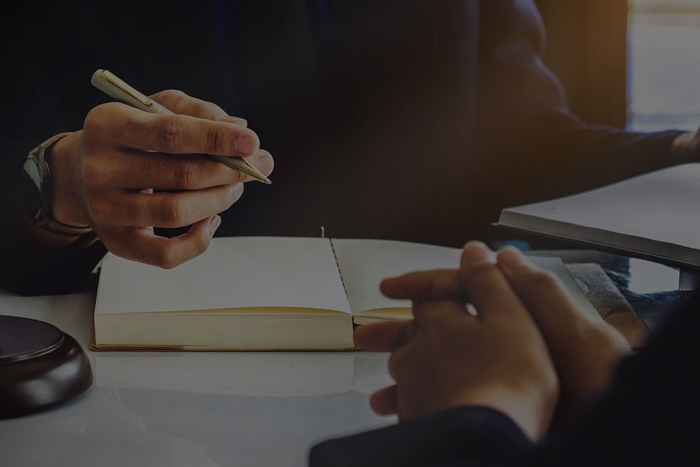 Opłaty Zdjęcie przedstawiające proces notowania, na pierwszym planie widać złożone ręce jednej z osób, w tle wiać otwarty, gruby notatnik oraz dłoń trzymającą długopis złotego koloru.