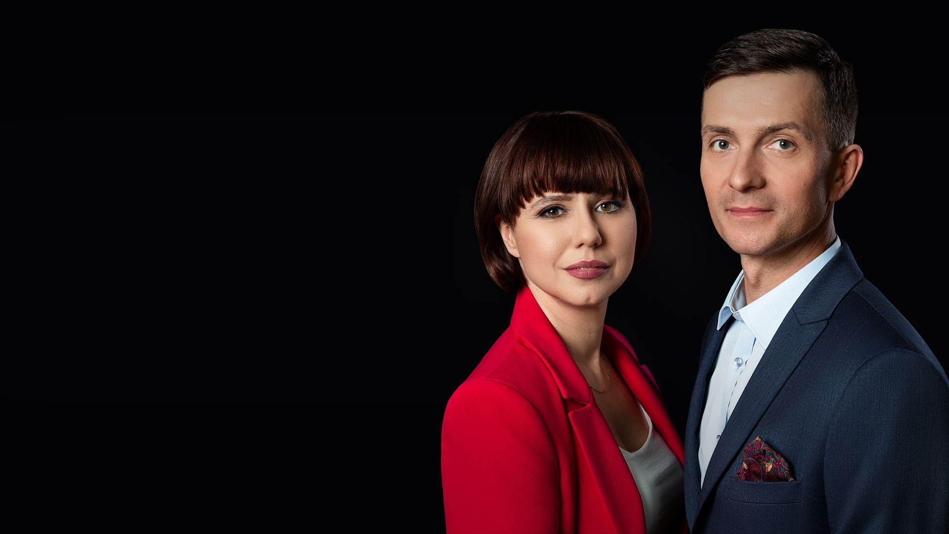 Zdjęcie przedstawiające stojących przy sobie Notariuszy Magdalenę Kułaj i Aleksandra Wasiaka. Ujęcie przedstawia Notariuszy od pasa w górę. Notariusz Magdalena Kułaj ubrana jest w czerwona marynarkę, białą podkoszulkę, Notariusz Aleksander Wasiak ubrany jest w granatową marynarkę, niebieską koszulę.