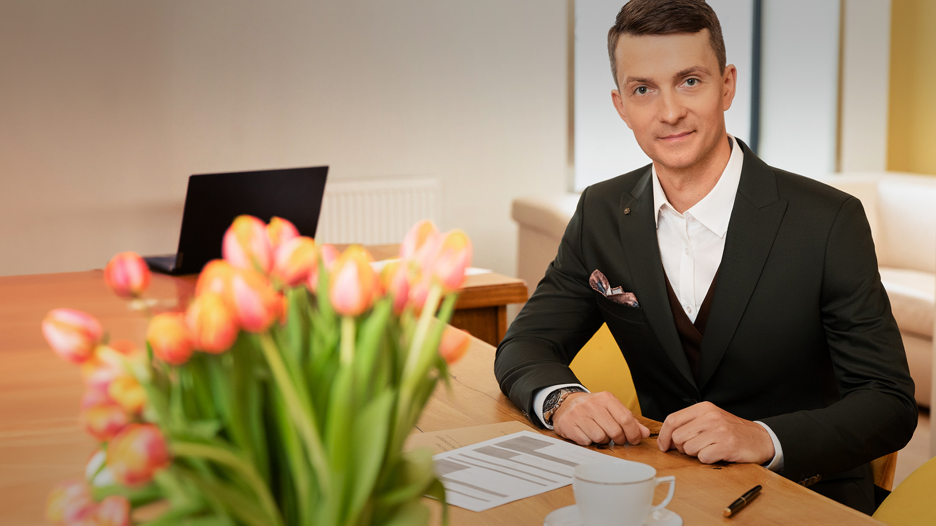 Zdjęcie siedzącego przy biurku Notariusza Aleksandra Wasiaka. Ubrany jest w grafitowy garnitur i białą koszulę. Przed nim widać dokument i czarny długopis oraz białą filiżankę z podstawką. Na pierwszym planie widać również bukiet żółtoczerwonych tulipanów. W tle widać, otwartego, czarnego laptopa.